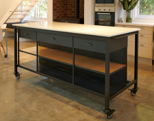 Load image into Gallery viewer, A dark grey metal kitchen island with a white quartz stone top, two drawers, and oak shelving. It has industrial castors and is displayed in a kitchen setting with stools in front of it.