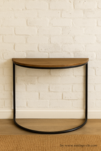 Load image into Gallery viewer, Wooden half-moon table with black metal frame against a white brick wall.
