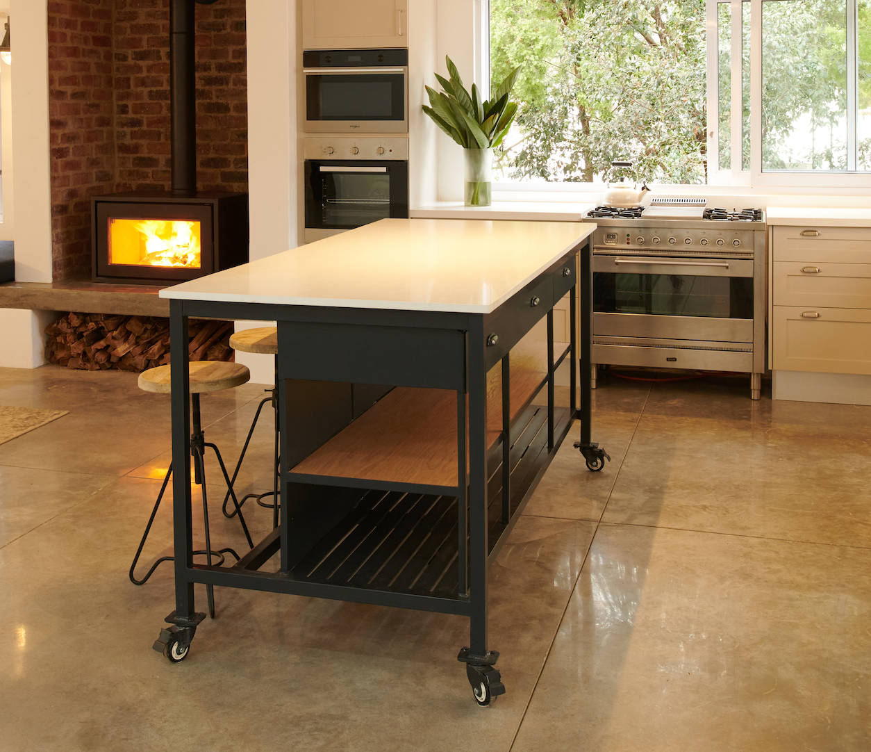 A dark grey metal kitchen island with a white quartz stone top, two drawers, and oak shelving. It has industrial castors and is displayed in a kitchen setting with stools in front of it.