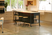 Load image into Gallery viewer, A dark grey metal kitchen island with a white quartz stone top, two drawers, and oak shelving. It has industrial castors and is displayed in a kitchen setting with stools in front of it.
