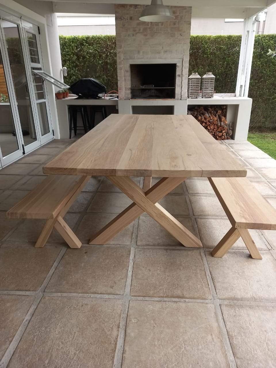A cross-legged oak table with benches, situated on a tiled outdoor patio area with a brick wall and a fireplace in the background.