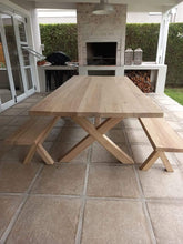 Load image into Gallery viewer, A cross-legged oak table with benches, situated on a tiled outdoor patio area with a brick wall and a fireplace in the background.
