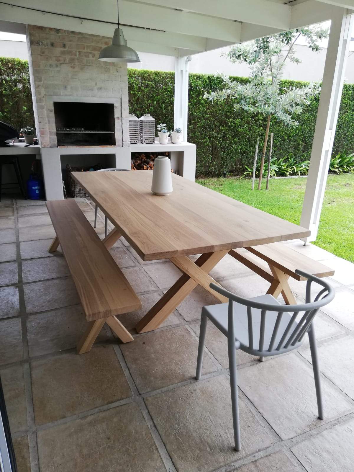 A cross-legged oak table with benches, situated on a tiled outdoor patio area with a brick wall and a fireplace in the background.