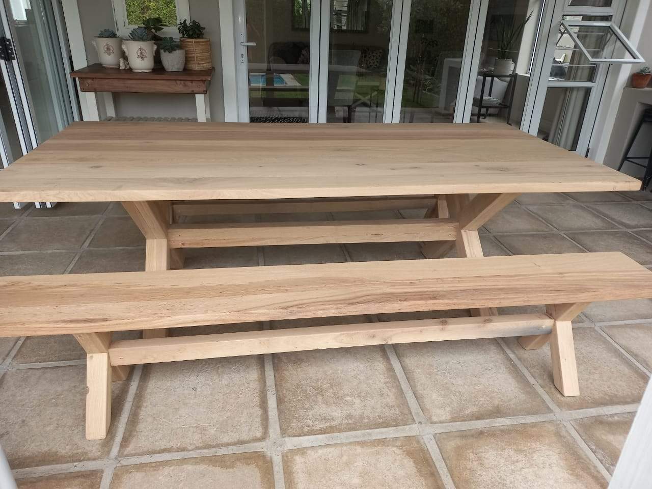 A cross-legged oak table with benches, situated on a tiled outdoor patio area with a brick wall and a fireplace in the background.