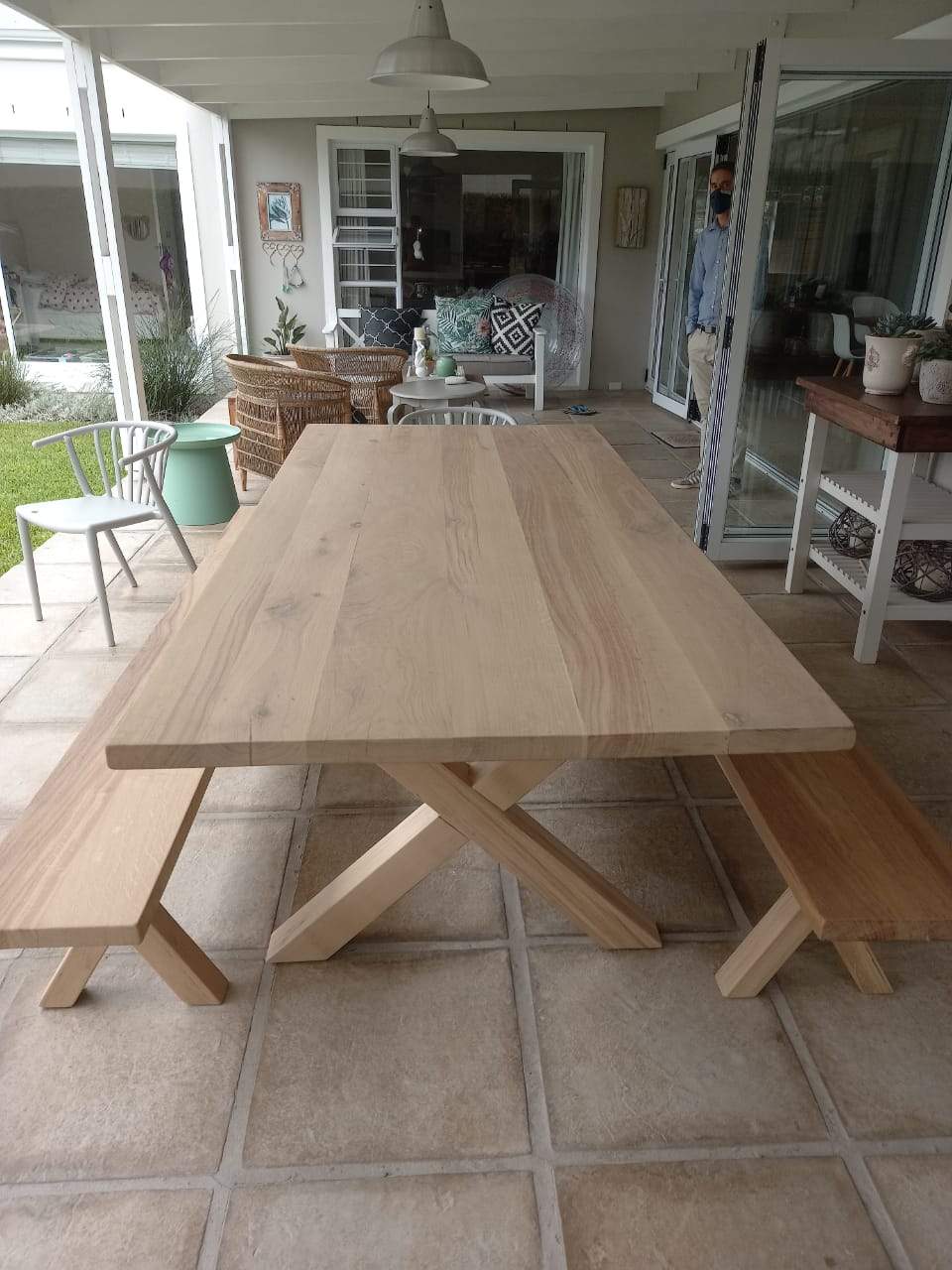 A cross-legged oak table with benches, situated on a tiled outdoor patio area with a brick wall and a fireplace in the background.
