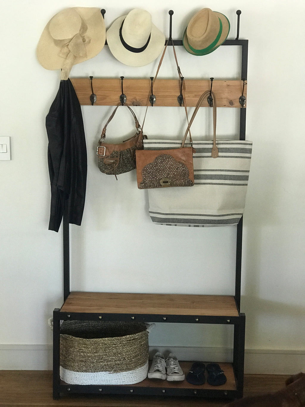 A black metal coat stand with a wooden shelf below, holding various items including hats, bags, and shoes.